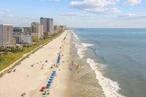 A beach with a row of blue umbrellas, a sandy shore, and buildings in the background. - Luxury Oceanfront 3BR w/ Hot Tub & Stunning View (Myrtle Beach)
