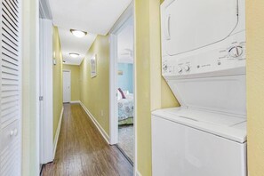A laundry room with a front-loading washer and dryer, a wooden floor, and a hallway leading to a bedroom with a bed. - Luxury Oceanfront 3BR w/ Hot Tub & Stunning View (Myrtle Beach)