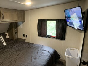 A bedroom with a bed, a television mounted on the wall, a window with curtains, and a trash bin.