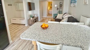 A modern kitchen with a granite countertop and a bowl of fruit. There is a dining area with a white table and chairs. The room has a neutral color scheme with light wood flooring.