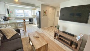 A modern living room with a sofa, coffee table, and TV stand. A kitchen is visible in the background with a refrigerator and cabinets.
