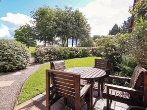 A wooden outdoor seating area with a table and chairs, surrounded by well-maintained hedges and trees.