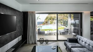 A modern living room with a large flat-screen TV, a grey sofa, and a glass door leading to a pool area with outdoor furniture.