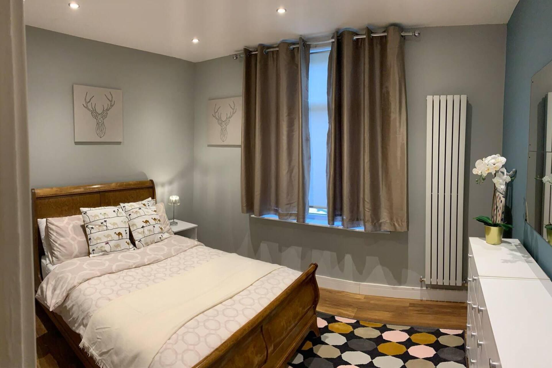 A bedroom with a wooden bed, a bedside table, a window with curtains, a radiator, and a mirror.