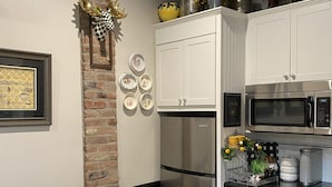 A kitchen with white cabinets, a stainless steel refrigerator, a microwave, and a coffee maker. There is a brick wall, a dining table with chairs, and a decorative wall hanging. - 1890 Graystone Cottage behind Historic Home short walk to Downtown & Whiskey Row (Prescott)