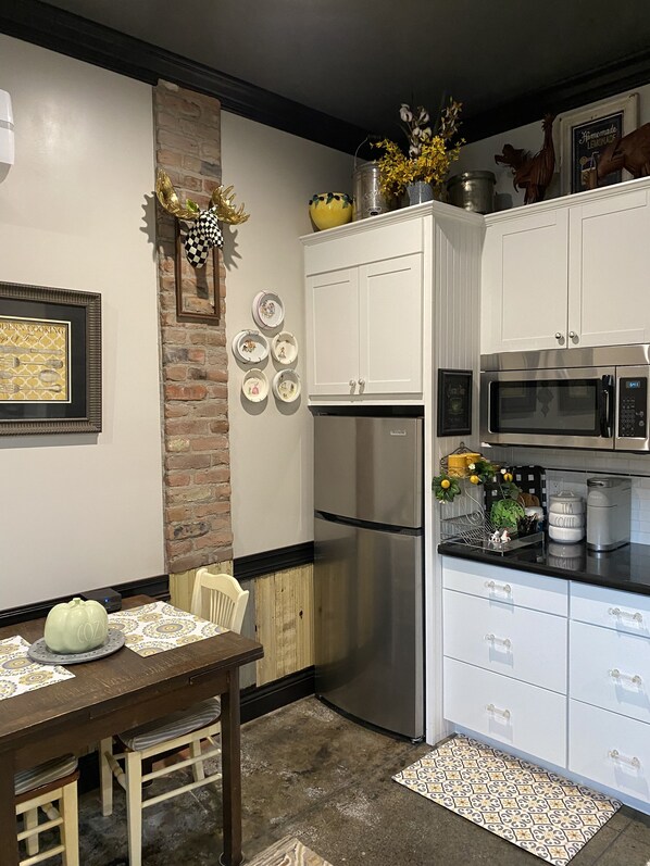 Una cocina con gabinetes blancos, un refrigerador de acero inoxidable, un microondas y una cafetera. Se observa una pared de ladrillo, una mesa de comedor con sillas y un adorno colgante.