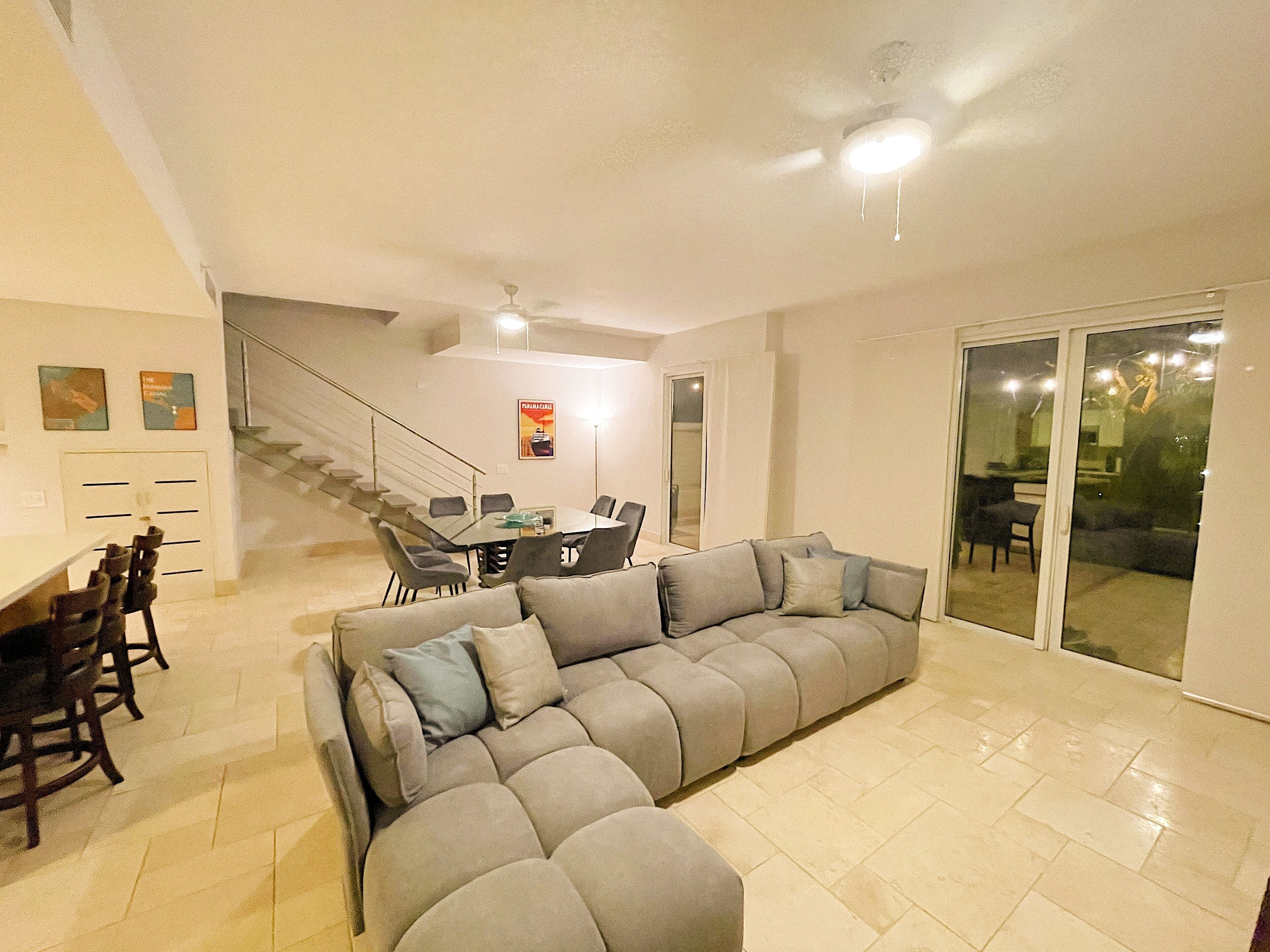 A spacious living area with a large gray sectional sofa, a dining table with chairs, and a staircase leading to an upper level.