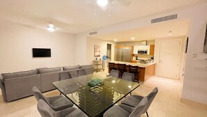 A modern living room with a glass dining table, grey sofas, and a kitchen in the background.
