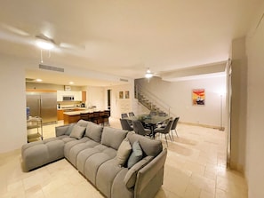 A modern living room with a sectional sofa, a dining table with chairs, a kitchen area with a refrigerator and microwave, and a staircase leading to another level. - Steps from Beach Club, Pools, Restaurants, Shopping and the Pacific!! (El Chirú)