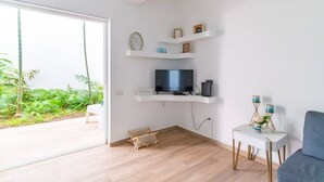 Ein modernes Wohnzimmer mit Fernseher auf weißem Regal, einem kleinen Beistelltisch mit Dekoration und Blick auf einen üppigen Garten durch eine Glasdoor.