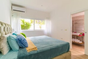A bedroom with a bed, a window with curtains, a bathroom with a sink and towels, and a wooden door.