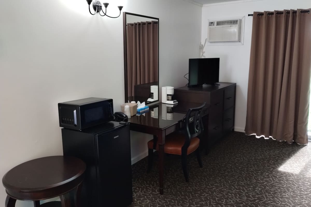 Habitaci ón de hotel con escritorio, silla, refrigerador pequeño, microondas, televisor, espejo, cafetera y ventana con cortinas.