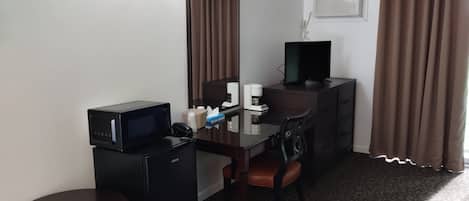 Habitación de hotel con escritorio, silla, refrigerador pequeño, microondas, televisor, espejo, cafetera y ventana con cortinas.