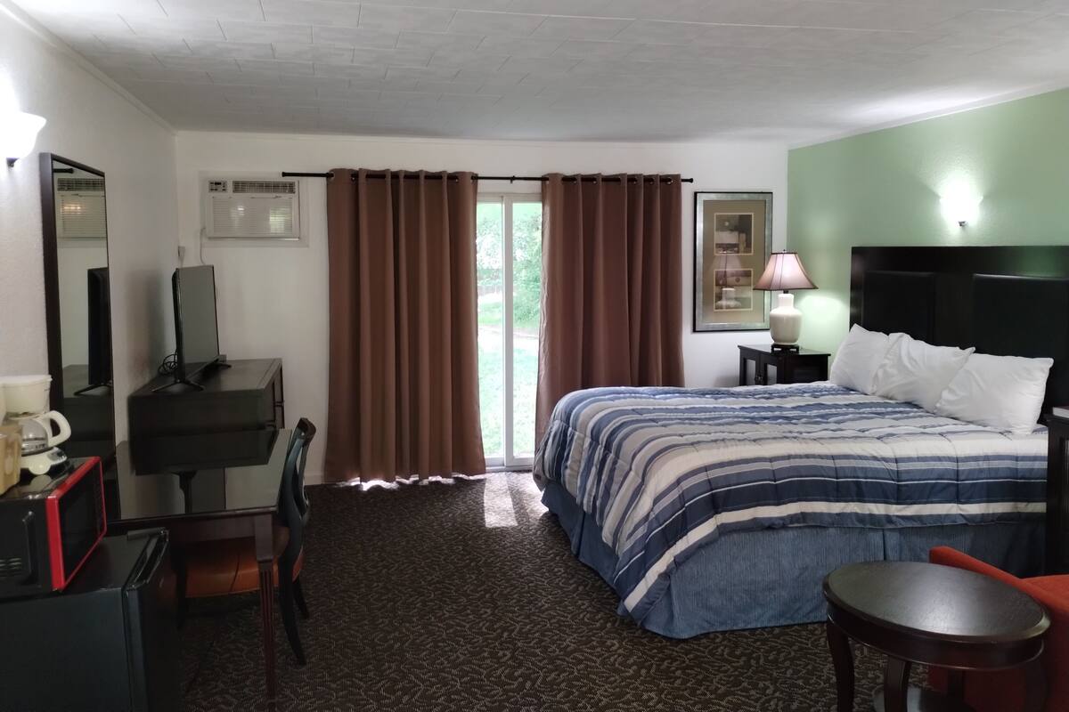 Habitación de hotel con una cama, un escritorio con computadora, una silla, una mesita y una ventana con cortinas.