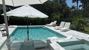 Une piscine rectangulaire à l’eau claire, entourée de chaises longues blanches et d’un parasol blanc.