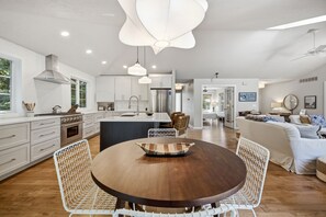 Een moderne keuken met witte kasten, een centraal eiland en een eetgedeelte met een ronde tafel en rieten stoelen.