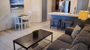 A modern living room with a sofa, a coffee table, and a dining area with a table and chairs. There is a kitchen area with a refrigerator and a bar counter. - Beachfront Lower Floor Luxury Condo (Puerto Peñasco)