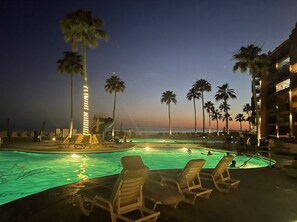 Zona de piscina con sillones reclinables, palmeras y un edificio con balcones que dan a la piscina.
