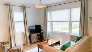 Een woonkamer met een bank, poef, salontafel, televisie en grote ramen.