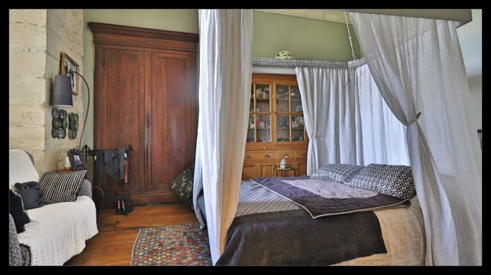 A bedroom with a wooden wardrobe, a bed with a canopy, and a small cabinet with glass doors.