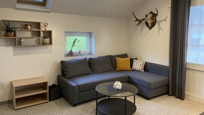 Ett modernt vardagsrum med en grå hörnsoffa, ett litet runt soffbord och en väggmonterad TV. Det finns hyllor med dekorativa föremål och ett stort fönster med gardiner.