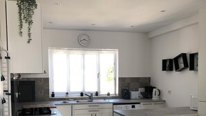 Eine moderne Küche mit weißen Schränken, einem Einbauherd, einer Spüle und einem Geschirrspüler. Ein Fenster bietet Blick auf viel Grün, und an der Wand hängt eine Uhr.