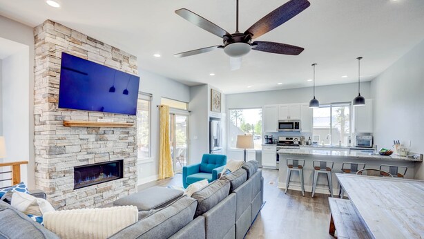 A modern living room with a stone fireplace, a large flat-screen TV, a ceiling fan, and a kitchen area with bar stools.