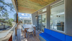 A covered patio with a wooden deck, a dining table with chairs, a blue cushioned seating area, and a view of a mountain range.
