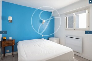 A bedroom with a blue wall, a white bed, a bedside table with a lamp, and a window with a view.