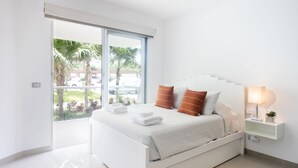 Une chambre moderne avec un lit blanc, une table de chevet avec une lampe et une vue sur des palmiers, visible à travers une porte coulissante.