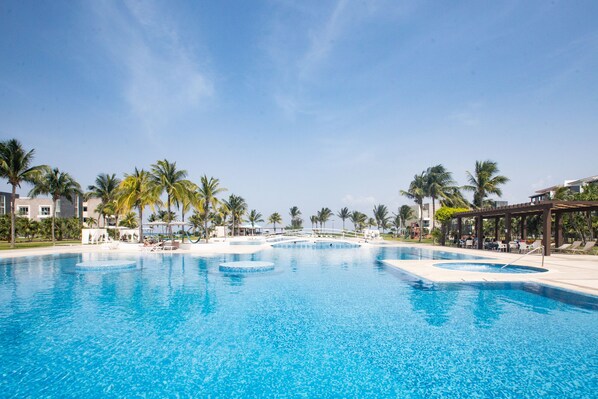 A large outdoor swimming pool surrounded by palm trees and lounge chairs, with a clear blue sky above.
