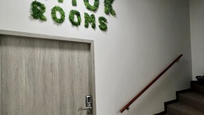 Una escalera con barandilla, una puerta de madera con un ojo de cerradura y una pared decorada con follaje verde que forma el texto "LIMOK ROOMS".
