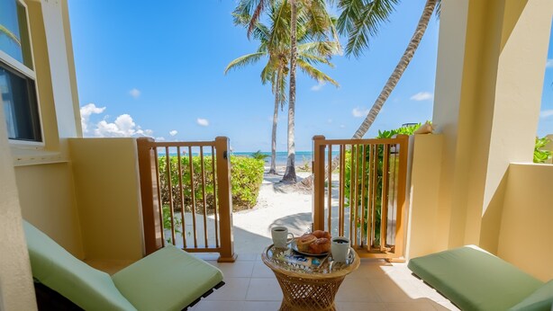 Een balkon met twee groene lounge fauteuils, een rieten tafeltje met een bord eten en twee kopjes, en uitzicht op het strand met palmbomen.