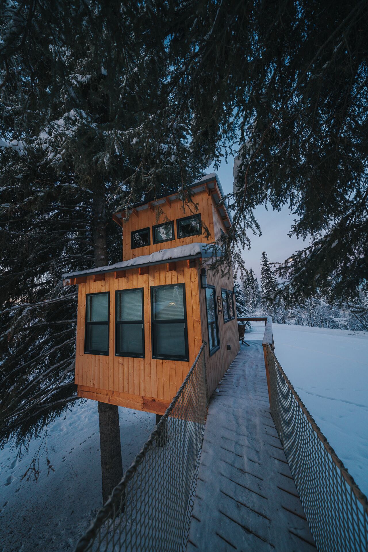 Une cabane en bois avec une terrasse en bois qui y mène, entourée d’arbres couverts de neige.