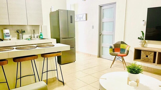 Moderni keittiö, jossa on jääkaappi, baaritiski ja baarijakkarat. Huoneen keskellä on pyöreä pöytä, jonka päällä on kasvi.