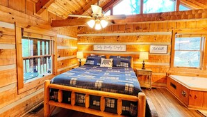 Une chambre dans une cabane en bois, avec un lit, deux fenêtres, un ventilateur de plafond et un panneau accroché au mur.