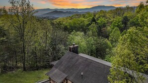Une cabane au toit en pente, entourée d’une forêt dense et avec des montagnes en arrière-plan.