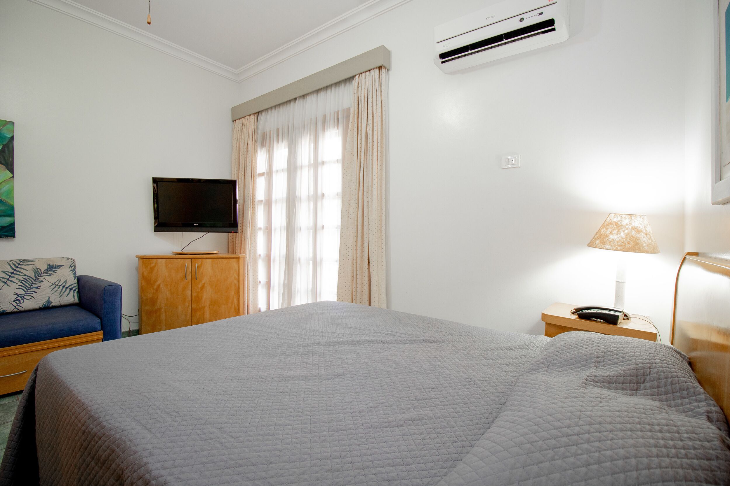 Une chambre d’hôtel avec un grand lit, un téléviseur fixé au mur, une table de chevet en bois, une lampe et une fenêtre avec des rideaux.