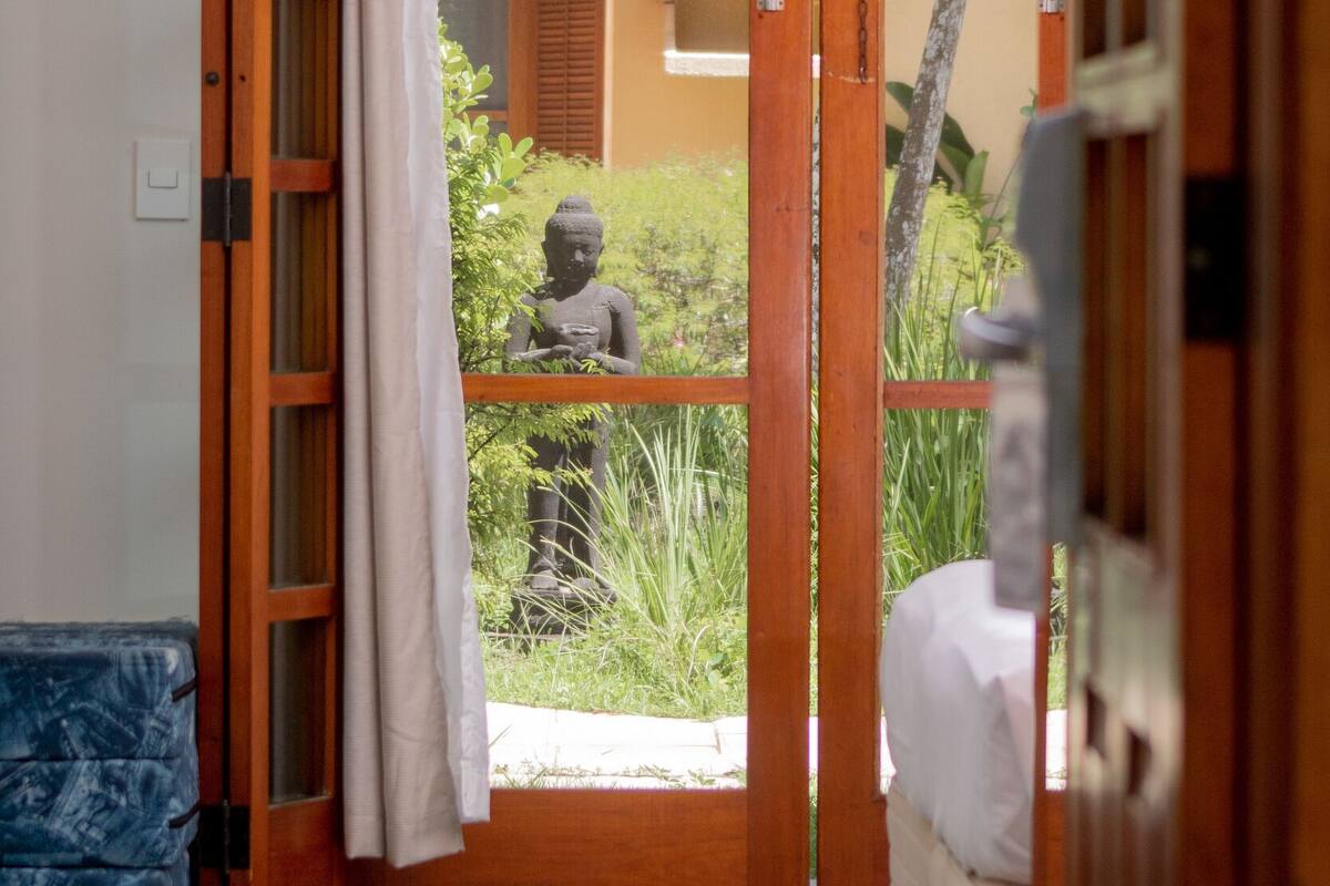 A room with a bed, a blue sofa, and a wooden door leading to a balcony with a statue and plants.