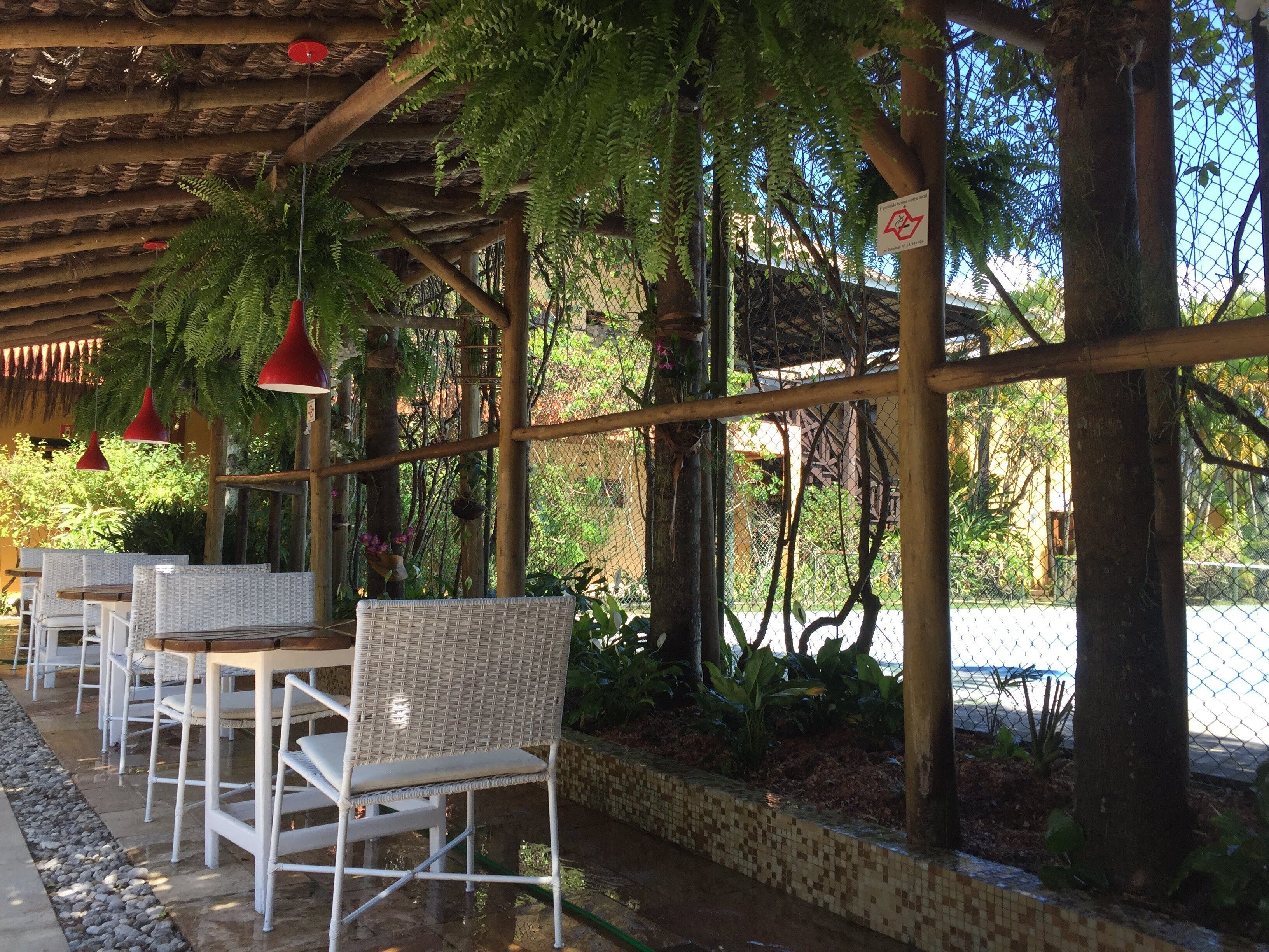 An outdoor seating area with wicker chairs and tables, a wooden pergola, and a chain-link fence with greenery.