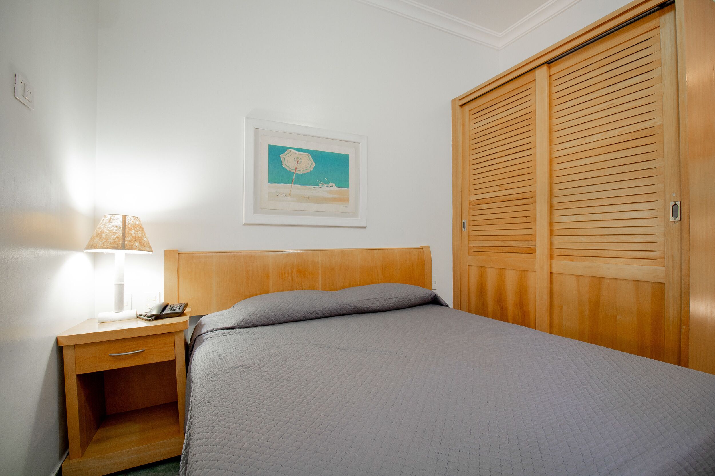 A neatly made bed with a grey quilt, a wooden nightstand with a lamp, and a wooden wardrobe.