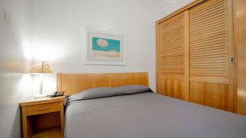 A neatly made bed with a grey quilt, a wooden nightstand with a lamp, and a wooden wardrobe.