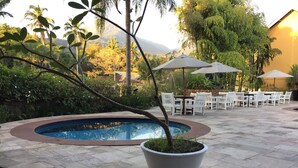 A pool area with a stone border, surrounded by white chairs and umbrellas, set against a backdrop of lush greenery and a mountain. - Ilha Flat Hotel (Ilhabela)