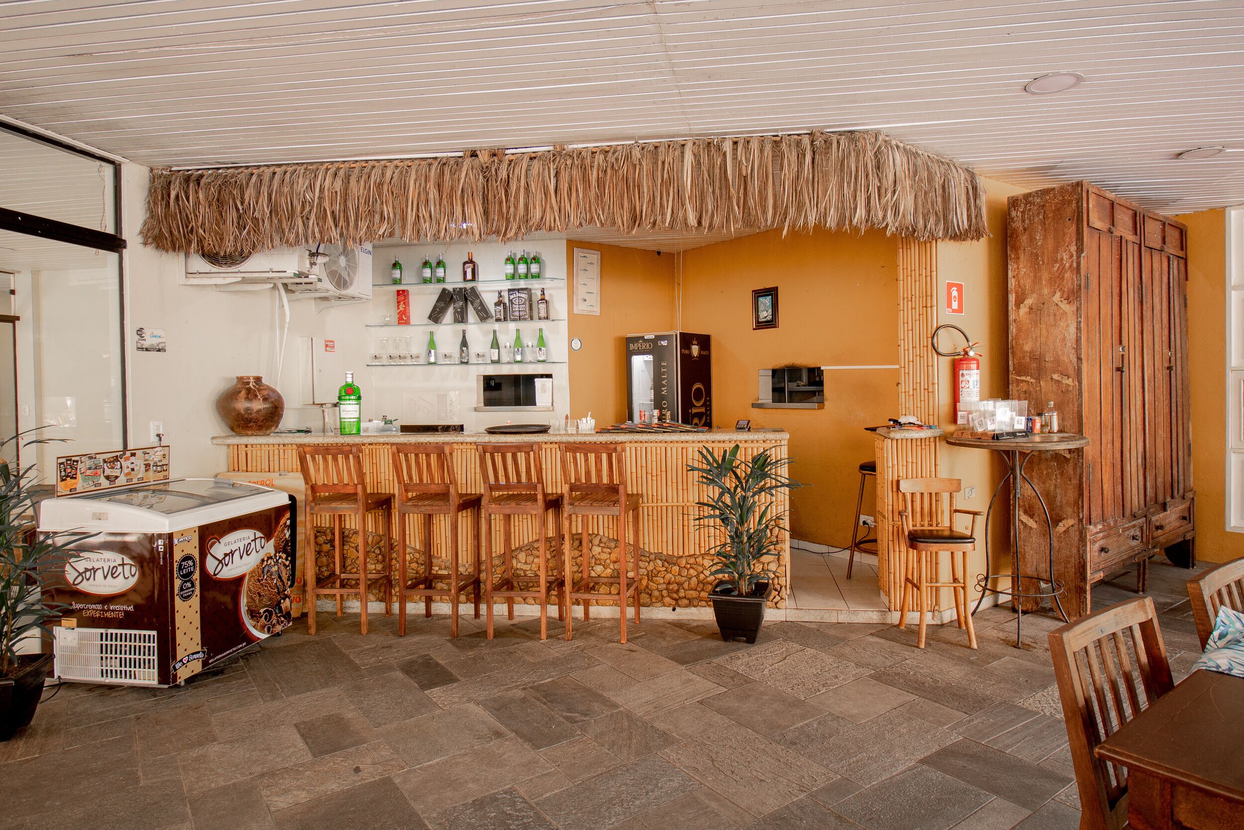 A bar area with a counter, stools, and a refrigerator. There are bottles on the wall and a fire extinguisher.