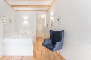 A hotel room with a bed, a chair, wooden flooring, and white walls.
