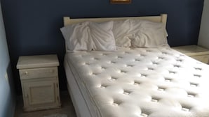 Um quarto com cama, mesa de cabeceira, armário pequeno, tapete fofo e um quadro na parede.
