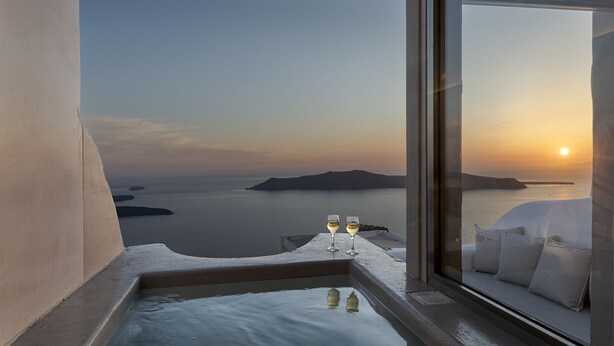 A balcony with a view of the sea and sunset, two glasses of wine, and a sofa with cushions. - Kaleidoscope Imerovigli Suites (Santorini)