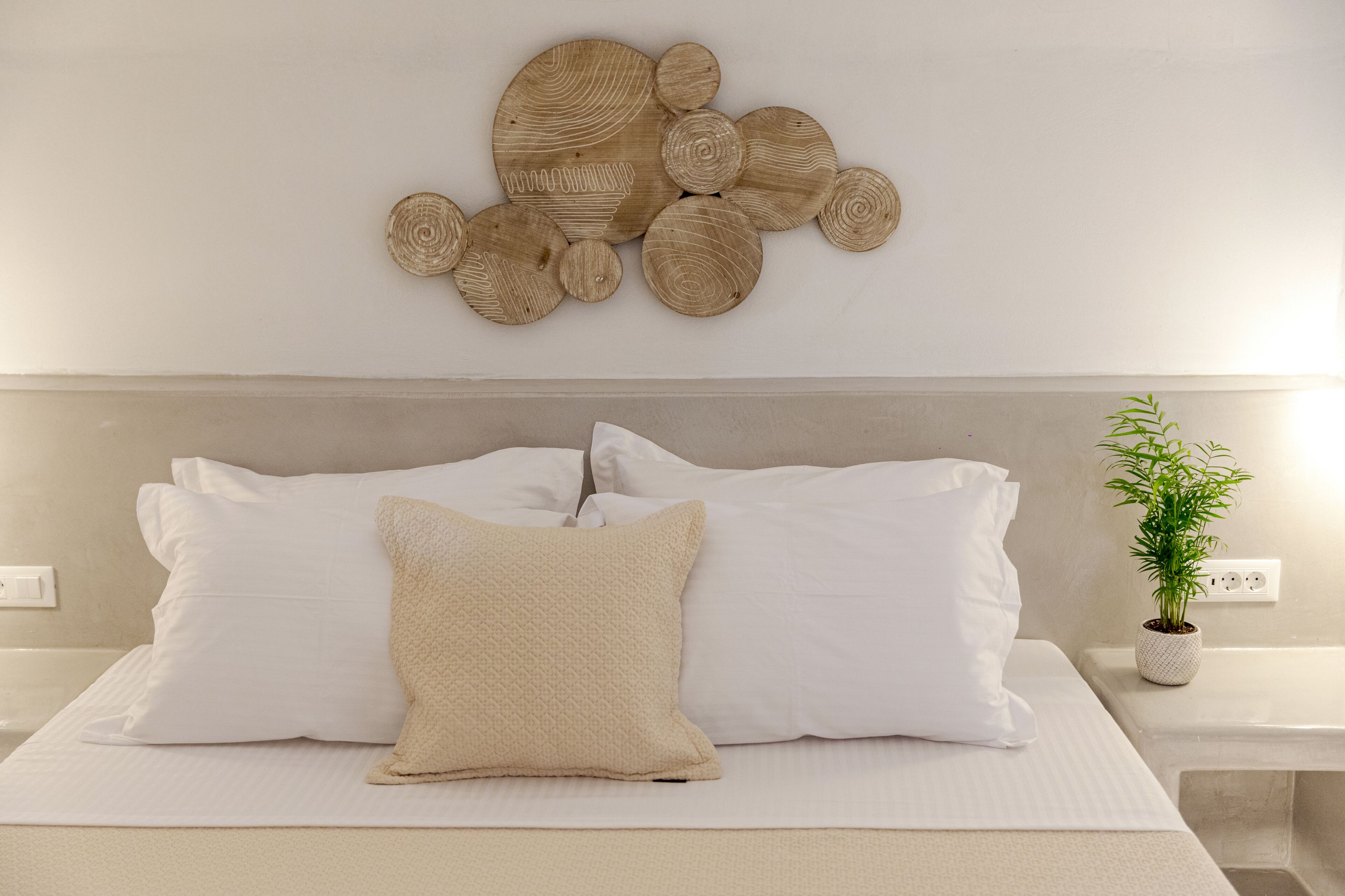 A neatly made bed with white linens and a textured beige pillow, a wall-mounted wooden art piece, and a small potted plant on a bedside table.