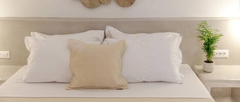 A neatly made bed with white linens and a textured beige pillow, a wall-mounted wooden art piece, and a small potted plant on a bedside table.