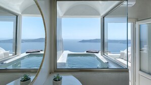 A room with a large window overlooking a body of water and hills. There is a small table with two potted plants and a glass-top table with a tray. - Kaleidoscope Imerovigli Suites (Santorini)